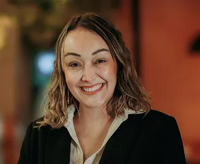A woman wearing a black blazer and white shirt smiles warmly at the camera