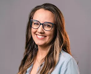 A woman wearing glasses and a blue shirt smiles warmly at the camera