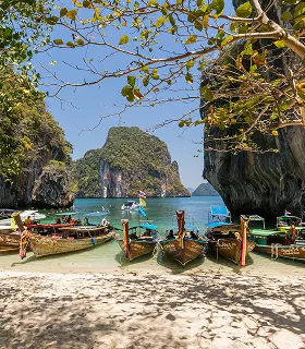 Longtail boats rest on a sandy beach under a leafy tree canopy