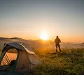 Man standing beside a tent, silhouetted against a vibrant sunset sky